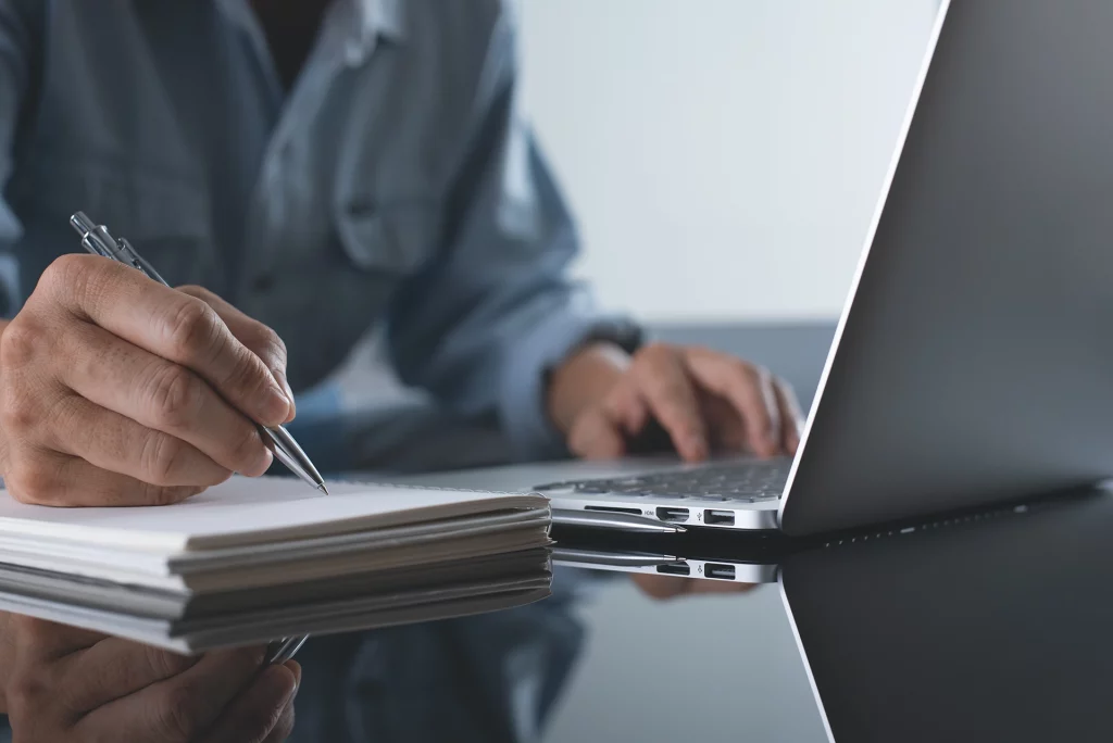 worker writing on notepad with laptop in front of them
